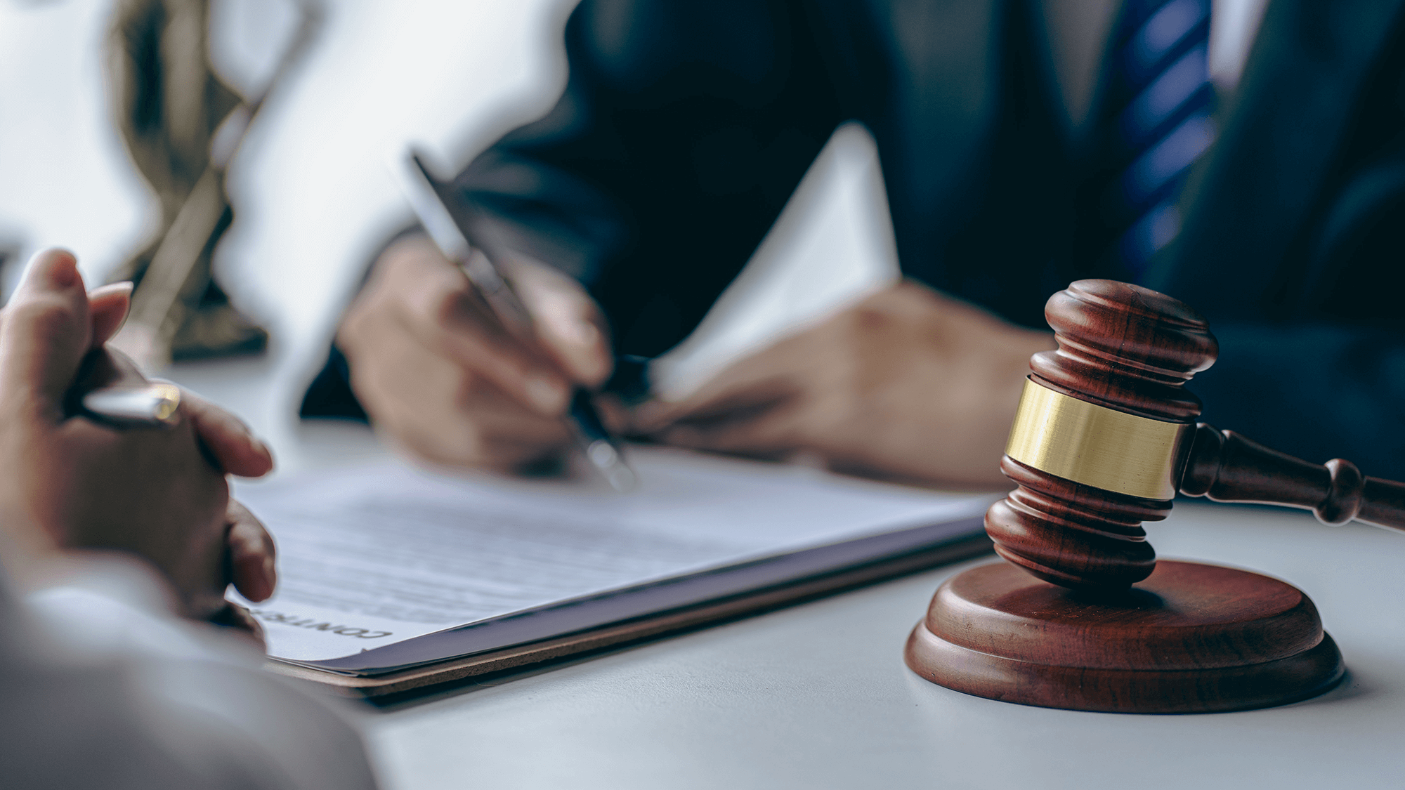A close-up of a gavel on a desk with a blurred hand writing on a legal document in the background, suggesting a courtroom setting.