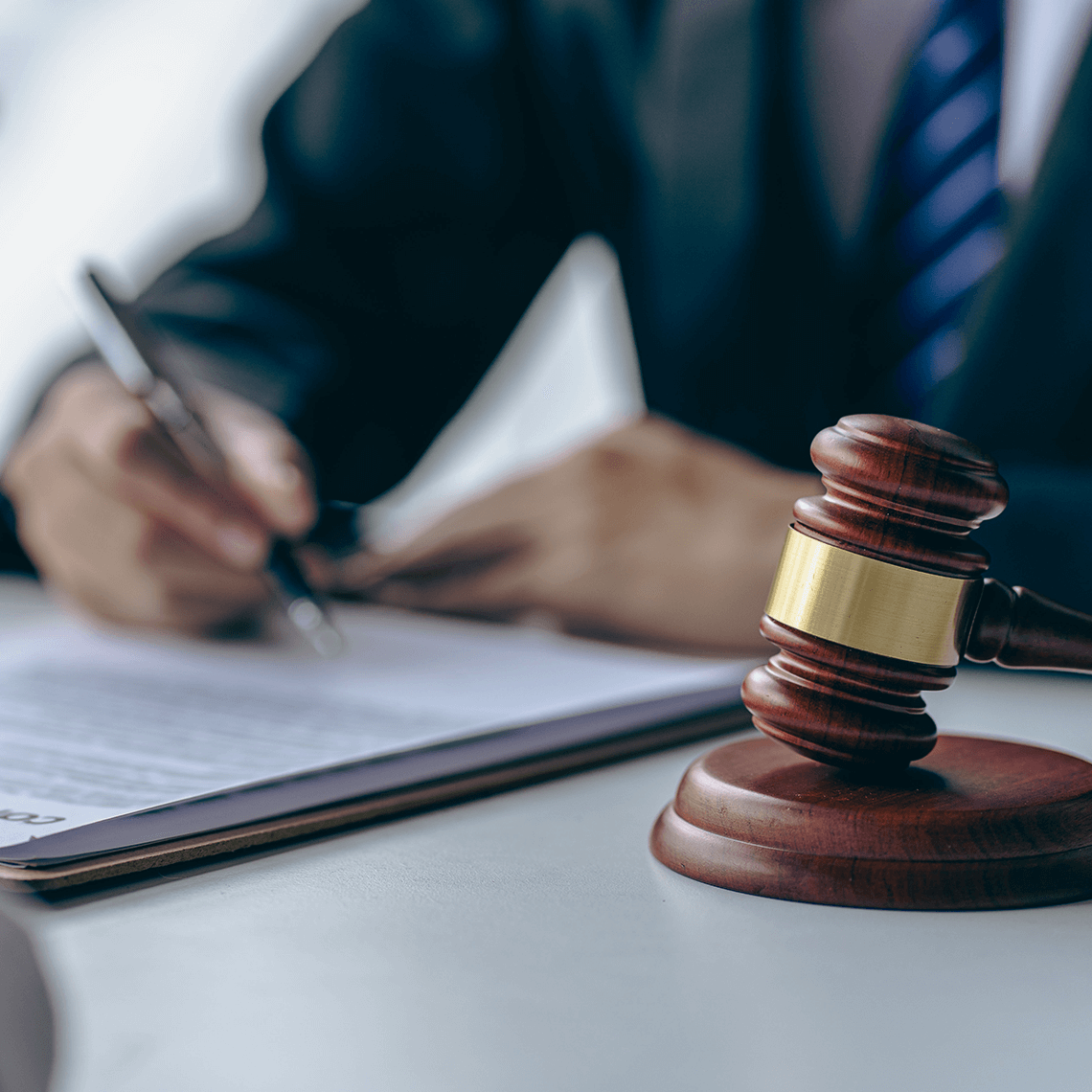 A close-up of a gavel on a desk with a blurred hand writing on a legal document in the background, suggesting a courtroom setting.
